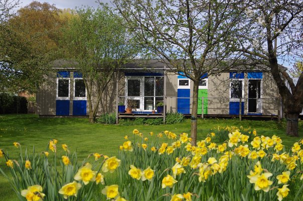 Betriebshof.jpg Ein flaches, modernes Garten- oder Bürogebäude mit farbigen Elementen hinter einer Wiese voller blühender gelber Narzissen.