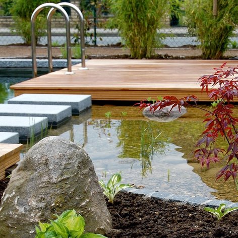 gartenteich-holzdeck.png Ein naturnah angelegter Gartenteich mit einer Holzterrasse und Trittsteinen im Wasser, umgeben von Beeten.