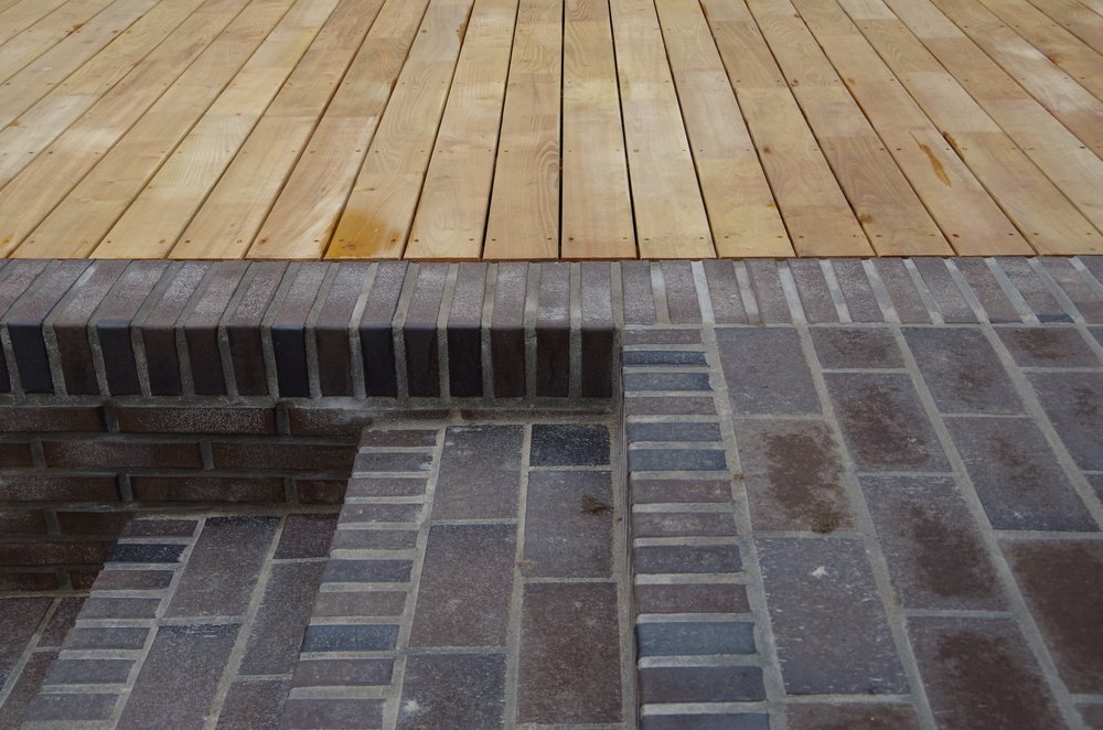 holzterrasse-klinker-treppe.png Eine Nahaufnahme zeigt den Übergang von hellen Holzdielen einer Terrasse zu einer gemauerten Treppe und Pflasterfläche aus dunklen Klinker- oder Backsteinen.