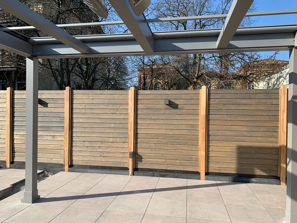 holzzaun-sichtschutz-terrasse.png Ein moderner Sichtschutzzaun aus horizontalen Holzlatten mit hellen Pfosten vor einer gepflasterten Terrasse mit Pergola-Struktur.