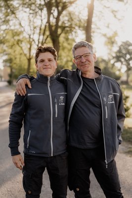 Anton_und_Gottfried_Bellin Zwei Männer stehen draußen im Park, lächeln in die Kamera und haben den Arm um die Schultern des jeweils anderen.
